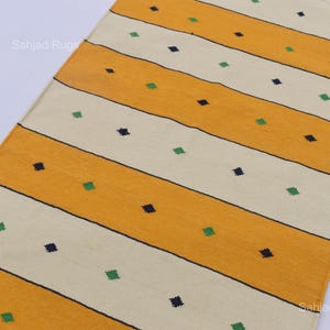 Puede incluir: Una alfombra rectangular con rayas amarillas y crema alternas. Peque&ntilde;os cuadrados azul oscuro y verde, espaciados uniformemente, salpican las rayas. La alfombra tiene un borde con flecos. El texto "Sahjad Rugs" es visible.