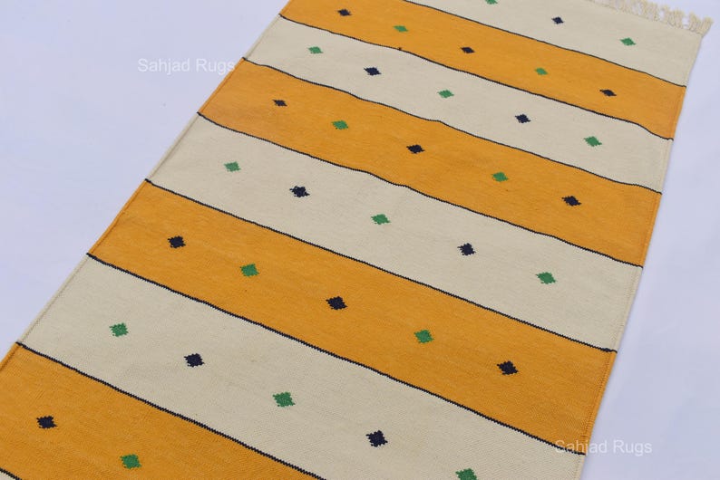 Puede incluir: Una alfombra rectangular con rayas amarillas y crema alternas. Peque&ntilde;os cuadrados azul oscuro y verde, espaciados uniformemente, salpican las rayas. La alfombra tiene un borde con flecos. El texto "Sahjad Rugs" es visible.