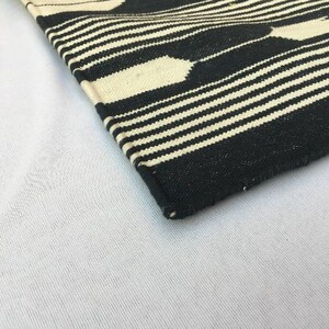 K&ouml;nnte beinhalten: Eine Ecke eines schwarz-wei&szlig; gewebten Textils. Das Design zeigt abwechselnd horizontale Streifen und geometrische Formen in Schwarz und Off-White. Das Textil ist wahrscheinlich ein Teppich oder ein dekorativer Stoff.