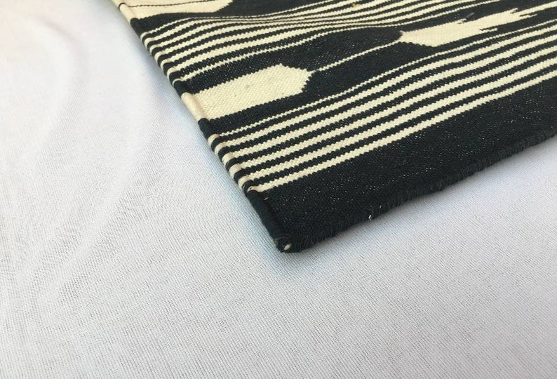 K&ouml;nnte beinhalten: Eine Ecke eines schwarz-wei&szlig; gewebten Textils. Das Design zeigt abwechselnd horizontale Streifen und geometrische Formen in Schwarz und Off-White. Das Textil ist wahrscheinlich ein Teppich oder ein dekorativer Stoff.