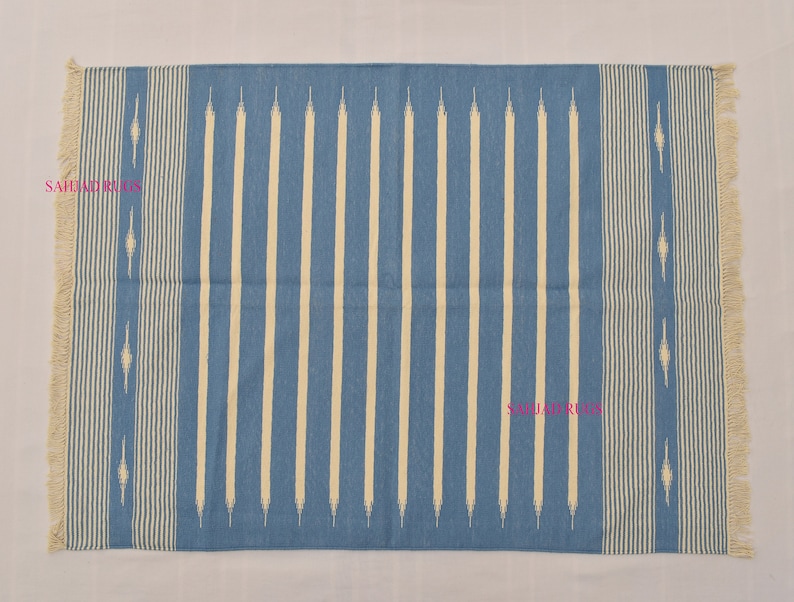May include: A rectangular blue rug with vertical cream-colored stripes and fringed edges. The rug features a repeating pattern of cream-colored diamond shapes along the sides. The rug is suitable for indoor use.