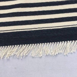 May include: A black and white striped rug with a white fringe. The rug is made of a woven material and has a simple design.