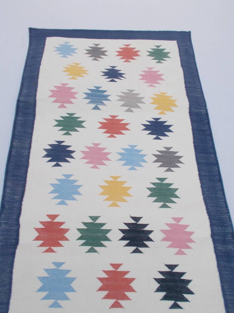 May include: A white runner rug with a blue border and a geometric pattern of colorful stars. The stars are in shades of blue, green, red, pink, yellow, and gray.