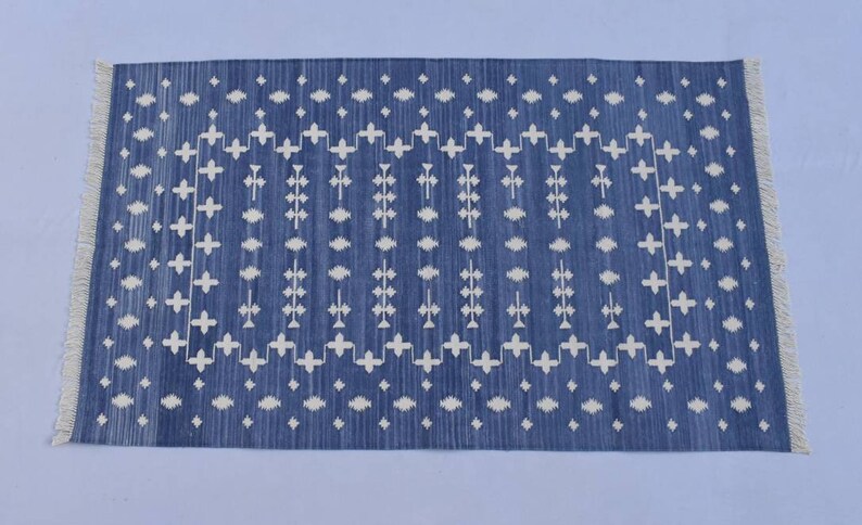 Puede incluir: Una alfombra tejida azul y blanca con un patr&oacute;n geom&eacute;trico. La alfombra tiene un borde de flecos.