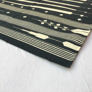 K&ouml;nnte beinhalten: Ein schwarz-cremefarbener gewebter Teppich mit geometrischem Muster. Der Teppich hat horizontale Streifen und rautenf&ouml;rmige Designs. Der Teppich besteht aus einem dicken, strapazierf&auml;higen Material und ist f&uuml;r den Innenbereich geeignet.