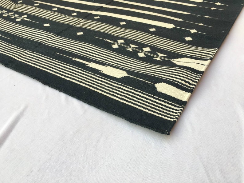 K&ouml;nnte beinhalten: Ein schwarz-cremefarbener gewebter Teppich mit geometrischem Muster. Der Teppich hat horizontale Streifen und rautenf&ouml;rmige Designs. Der Teppich besteht aus einem dicken, strapazierf&auml;higen Material und ist f&uuml;r den Innenbereich geeignet.