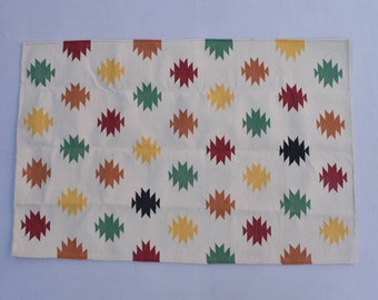 Puede incluir: Una alfombra blanca con un patrón geométrico en tonos de rojo, naranja, amarillo, verde y negro. El patrón está formado por formas de diamante que se repiten.
