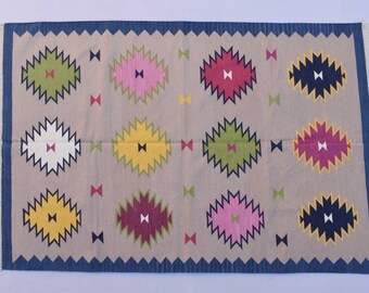 May include: A beige rug with a blue border and a repeating pattern of colorful diamond shapes. The rug has a fringe on the edges.