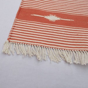 May include: A close-up of a woven rug with orange and white stripes and a fringe border. The rug has a geometric pattern in the center.