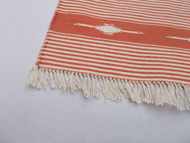 May include: A close-up of a woven rug with orange and white stripes and a fringe border. The rug has a geometric pattern in the center.