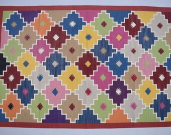 May include: A rectangular rug with a geometric pattern. The rug features a repeating diamond design in various colors, including blue, green, yellow, pink, and brown, set against a beige background. The border is a solid red, and the rug has fringed edges.