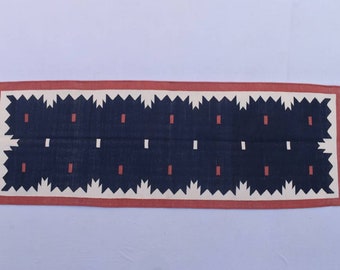May include: A navy blue and white runner rug with a geometric pattern. The rug has a red border and white fringe.
