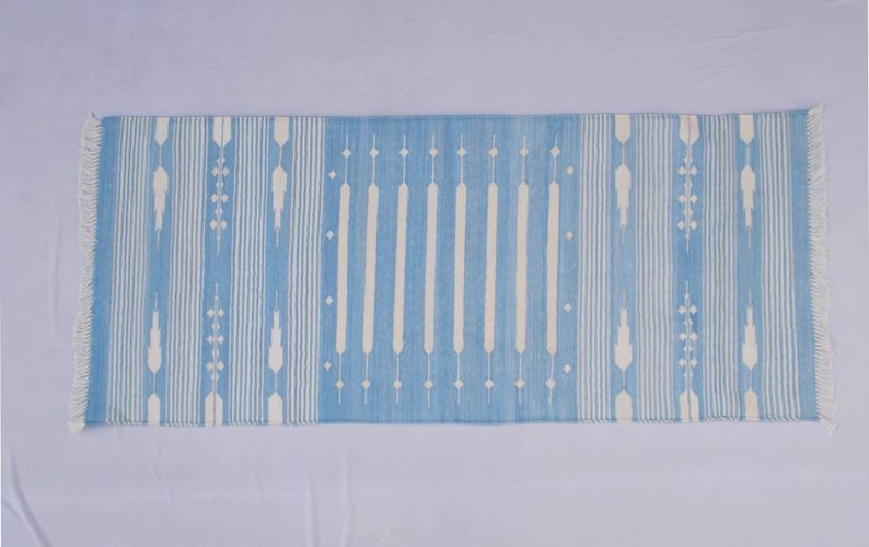 Puede incluir: Una alfombra a rayas azul y blanca con un patr&oacute;n geom&eacute;trico. La alfombra tiene flecos en ambos extremos.