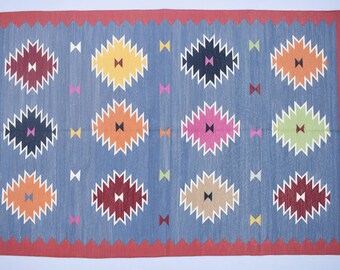 May include: A rectangular blue rug with a red border and fringed edges. The rug features a repeating pattern of colorful, stylized starburst shapes in various colors, including yellow, orange, and burgundy. Small bow tie shapes in multiple colors are scattered across the rug.