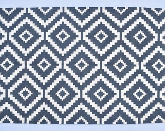 May include: A rectangular rug featuring a geometric diamond pattern in dark grey and white. The rug has a fringe on both short ends. This rug is suitable for living rooms, bedrooms, or entryways.