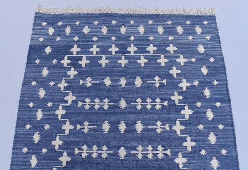 Puede incluir: Una alfombra azul y blanca con un dise&ntilde;o geom&eacute;trico. La alfombra tiene un borde de flecos.