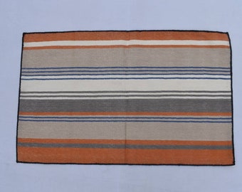 Puede incluir: Una alfombra rayada con un borde negro. La alfombra tiene rayas de color naranja, marrón, gris, blanco y azul.
