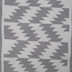 Op de afbeelding: Een wit en grijs geometrisch tapijt met een franje rand. Het tapijt heeft een chevron patroon.