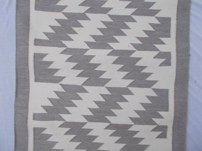 Op de afbeelding: Een wit en grijs geometrisch tapijt met een franje rand. Het tapijt heeft een chevron patroon.