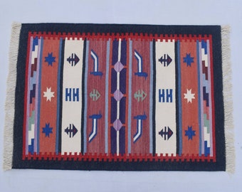 Puede incluir: Una alfombra colorida con un patrón geométrico en tonos de rojo, azul, blanco y negro. La alfombra tiene un borde con flecos y presenta un diseño repetitivo de estrellas, flechas y figuras estilizadas.
