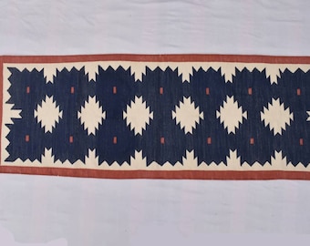 May include: A long, narrow rug with a geometric pattern in shades of blue, white, and red. The rug has a fringe on both ends.