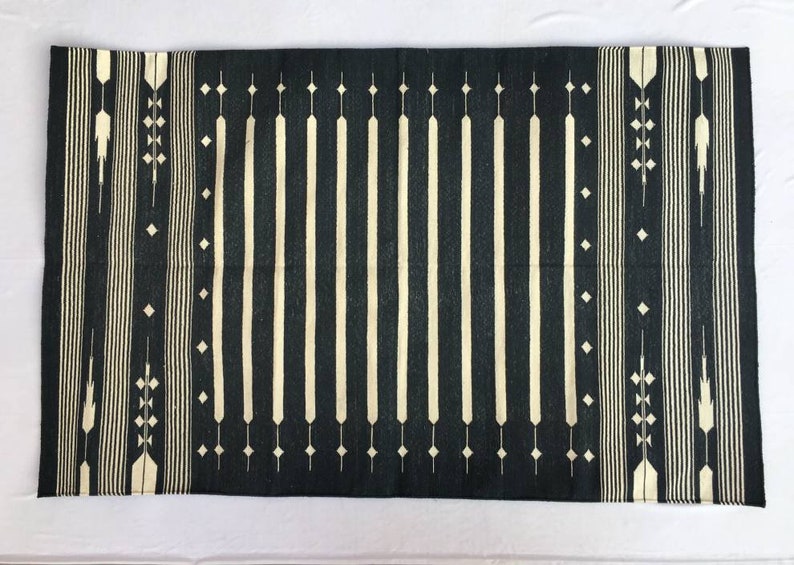 K&ouml;nnte beinhalten: Ein rechteckiger, dunkelblauer Teppich mit einem cremefarbenen geometrischen Muster. Das Design zeigt vertikale Streifen in der Mitte und dekorative Elemente an den Seiten, wodurch ein symmetrisches und optisch ansprechendes Textil entsteht.