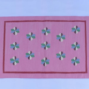 Könnte beinhalten: Rechteckiger rosa Teppich mit einem Blumenmuster in Blau-, Grün- und Weißtönen. Der Teppich hat einen dunkelroten Rand und Fransen an den kurzen Enden. Ein dekorativer Bodenbelag.