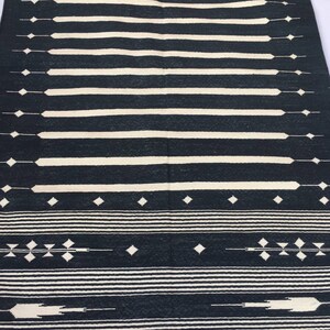 K&ouml;nnte beinhalten: Ein schwarz-wei&szlig;er gewebter Teppich mit geometrischem Muster. Der Teppich hat horizontale wei&szlig;e Streifen und rautenf&ouml;rmige Akzente. Das Design ist symmetrisch und der Teppich ist wahrscheinlich aus Naturfasern gefertigt.