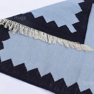May include: A light blue and black geometric patterned rug with a fringed edge. The rug has a repeating zigzag design in black along the border. The rug is made of a woven material.