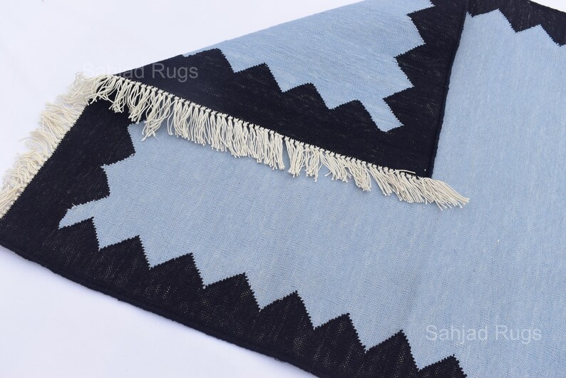 May include: A light blue and black geometric patterned rug with a fringed edge. The rug has a repeating zigzag design in black along the border. The rug is made of a woven material.