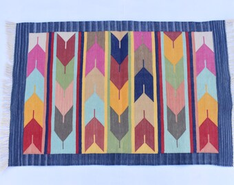 Puede incluir: Una alfombra rectangular con borde azul marino y bordes con flecos. La alfombra presenta un patrón central de filas verticales de formas coloridas en forma de flecha en tonos de rojo, rosa, amarillo, verde y azul. La alfombra probablemente está hecha de tela tejida.