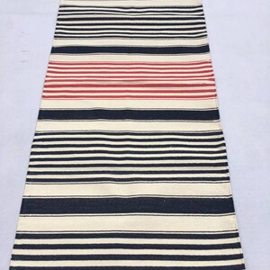 May include: A black and white striped rug with a red stripe in the middle. The rug has a fringe on both ends.