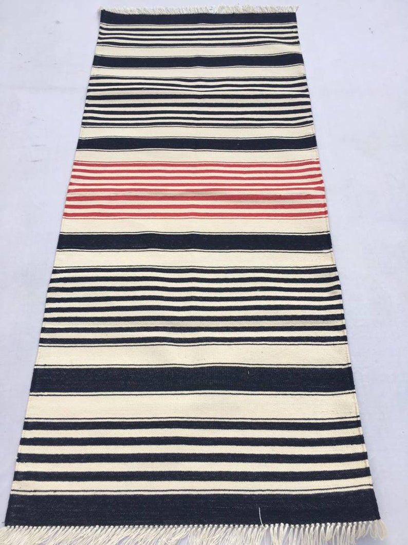 May include: A black and white striped rug with a red stripe in the middle. The rug has a fringe on both ends.