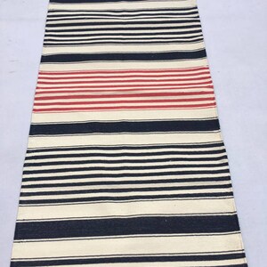May include: A long, narrow, cream-coloured rug with black and red stripes. The rug has a fringe on the top and bottom.