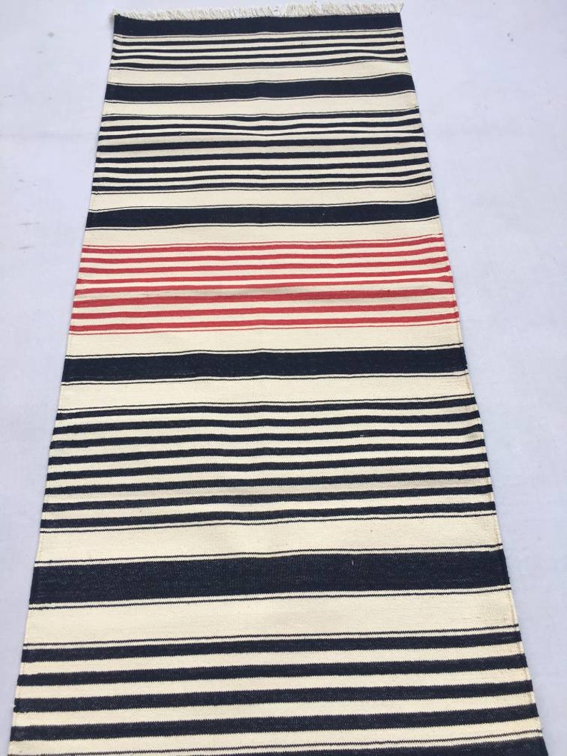 May include: A long, narrow, cream-coloured rug with black and red stripes. The rug has a fringe on the top and bottom.