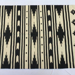 May include: Black and white geometric patterned rug with fringe on the edges. The rug has a tribal design with black and white stripes and geometric shapes.