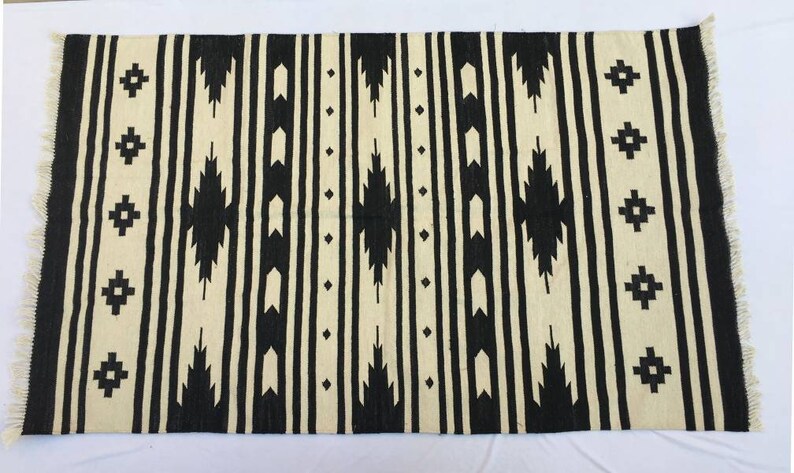 May include: Black and white geometric patterned rug with fringe on the edges. The rug has a tribal design with black and white stripes and geometric shapes.