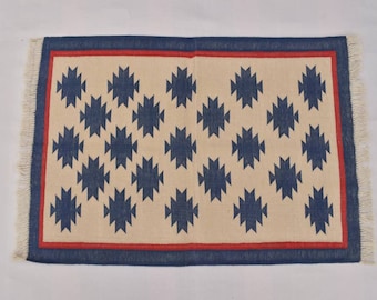 May include: A rectangular rug with a cream background and a blue geometric pattern. The rug has a red and blue border and white fringe.