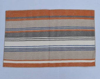Puede incluir: Una alfombra rayada con rayas naranjas, grises, blancas y azules. La alfombra está hecha de un material tejido y tiene una superficie ligeramente texturizada.