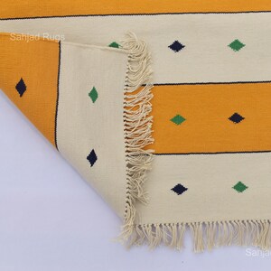 Puede incluir: Una alfombra rectangular plegada con un patr&oacute;n repetitivo de rayas horizontales en amarillo y blanco roto. El dise&ntilde;o incluye peque&ntilde;as formas de diamante en azul oscuro y verde. La alfombra tiene un borde con flecos.