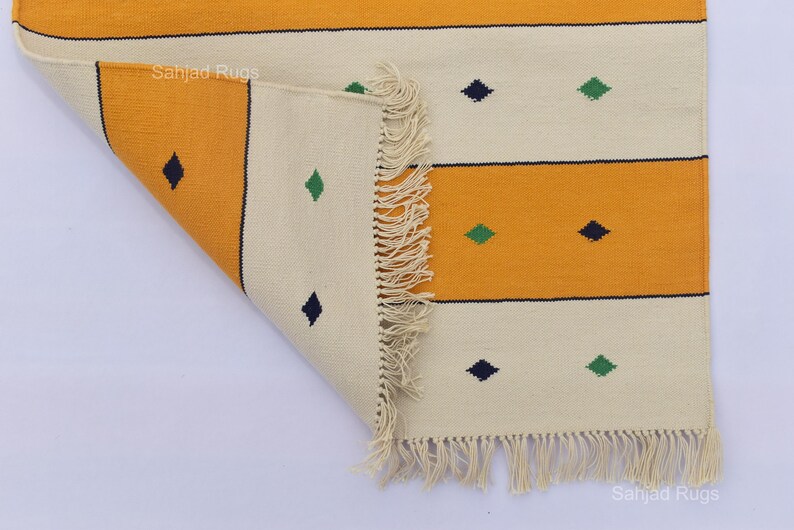 Puede incluir: Una alfombra rectangular plegada con un patr&oacute;n repetitivo de rayas horizontales en amarillo y blanco roto. El dise&ntilde;o incluye peque&ntilde;as formas de diamante en azul oscuro y verde. La alfombra tiene un borde con flecos.