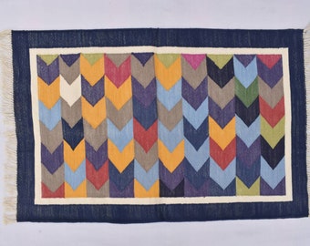 Puede incluir: Una alfombra colorida con un patrón de chevron en tonos de azul, amarillo, rojo, verde y morado. La alfombra tiene un borde azul oscuro y flecos blancos.