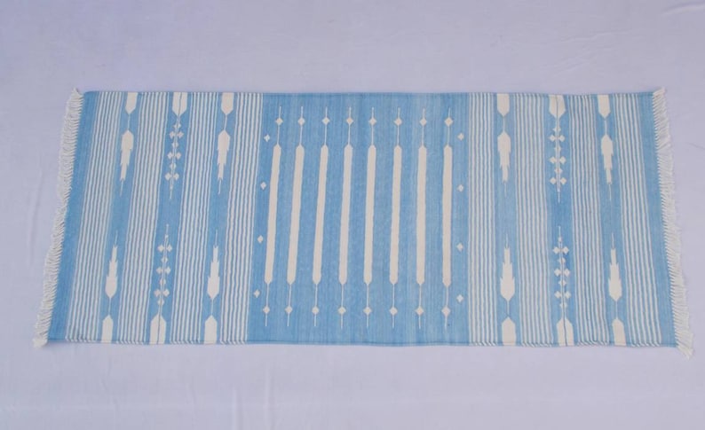 Puede incluir: Una alfombra de rayas azules y blancas con un patr&oacute;n geom&eacute;trico. La alfombra tiene flecos en ambos extremos.
