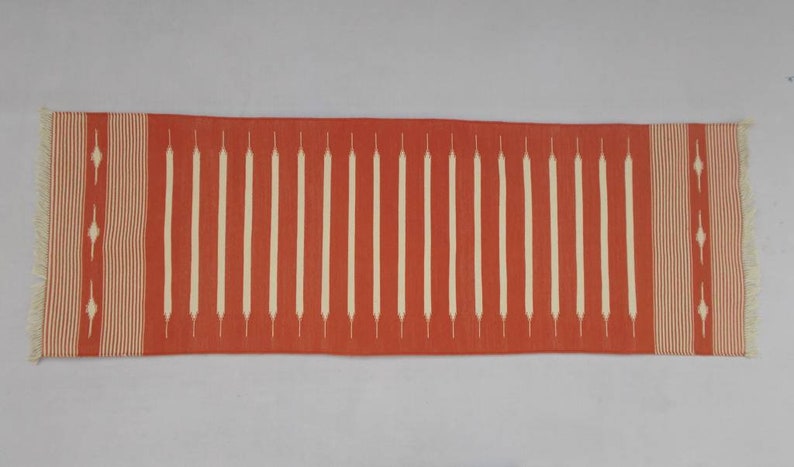 May include: A long, rectangular red runner rug with a white geometric pattern. The rug has fringe on both ends.