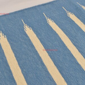 May include: Blue and cream-colored rug with a striped pattern. The rug features alternating bands of light blue and cream-colored stripes. The cream stripes have a textured, elongated triangular design. The rug is made of woven material.