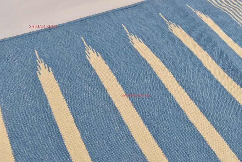 May include: Blue and cream-colored rug with a striped pattern. The rug features alternating bands of light blue and cream-colored stripes. The cream stripes have a textured, elongated triangular design. The rug is made of woven material.