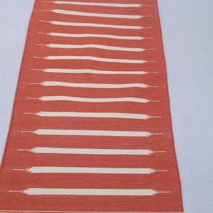 May include: A long, narrow, red and white striped rug. The rug has a woven texture and is made of cotton.