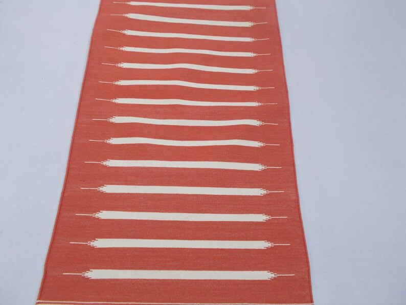 May include: A long, narrow, red and white striped rug. The rug has a woven texture and is made of cotton.