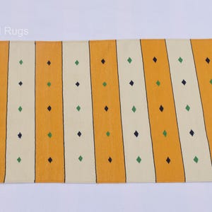 Puede incluir: Una alfombra rectangular con un patr&oacute;n de rayas. Rayas verticales alternas amarillas y blanquecinas se acent&uacute;an con filas de peque&ntilde;os rombos verdes y negros. La alfombra tiene bordes con flecos.
