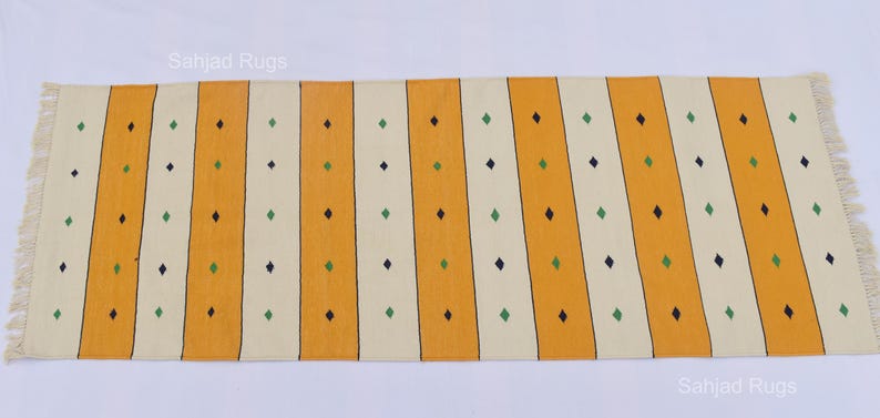 Puede incluir: Una alfombra rectangular con un patr&oacute;n de rayas. Rayas verticales alternas amarillas y blanquecinas se acent&uacute;an con filas de peque&ntilde;os rombos verdes y negros. La alfombra tiene bordes con flecos.